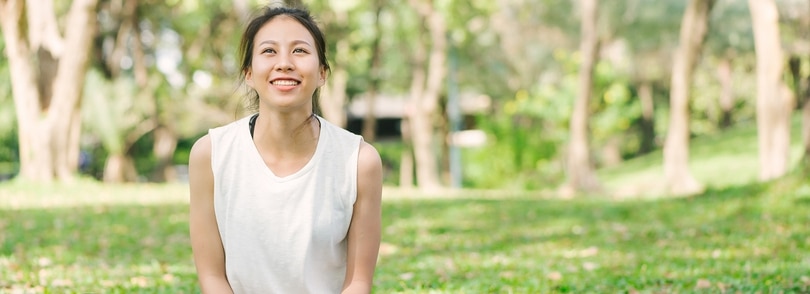 An Asian woman peacefully sitting on grass in a park, with beautiful trees in the background