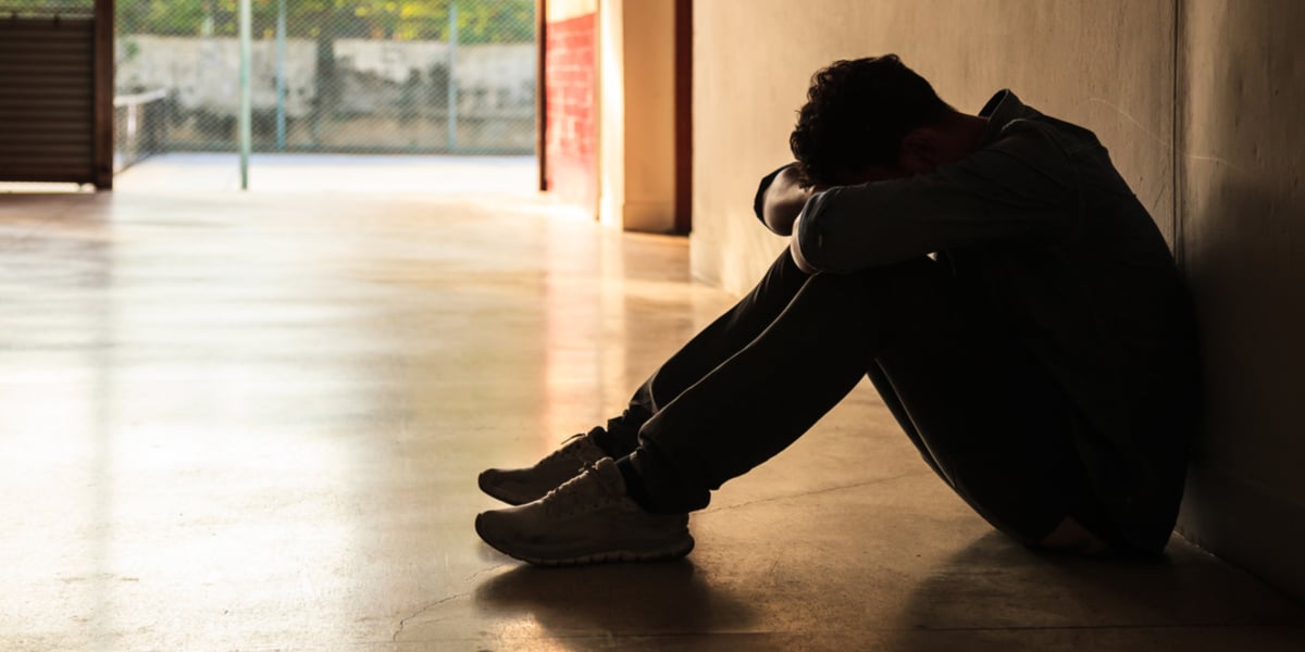 Person in shadow seated on the floor of a hallway, while resting his head on his folded arms resting on his knees