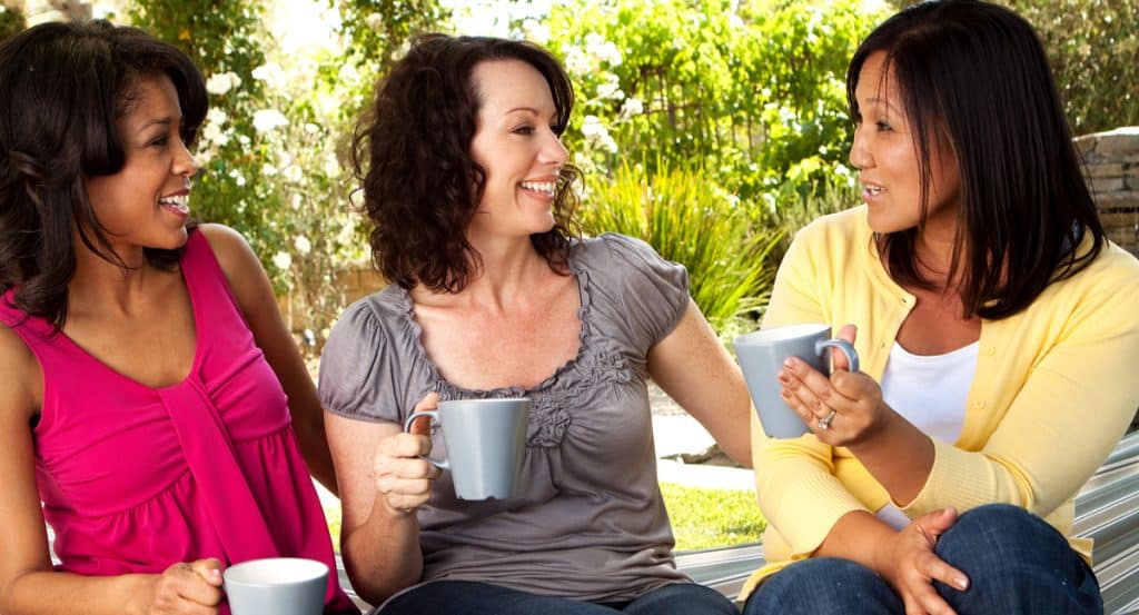 three women seated in conversation outside holding coffee cups