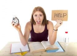 College woman at desk full of papers and work holding up a clock and a hand-written sign
