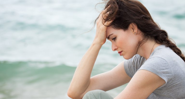 Woman seated near the ocean resting her forehead in one hand