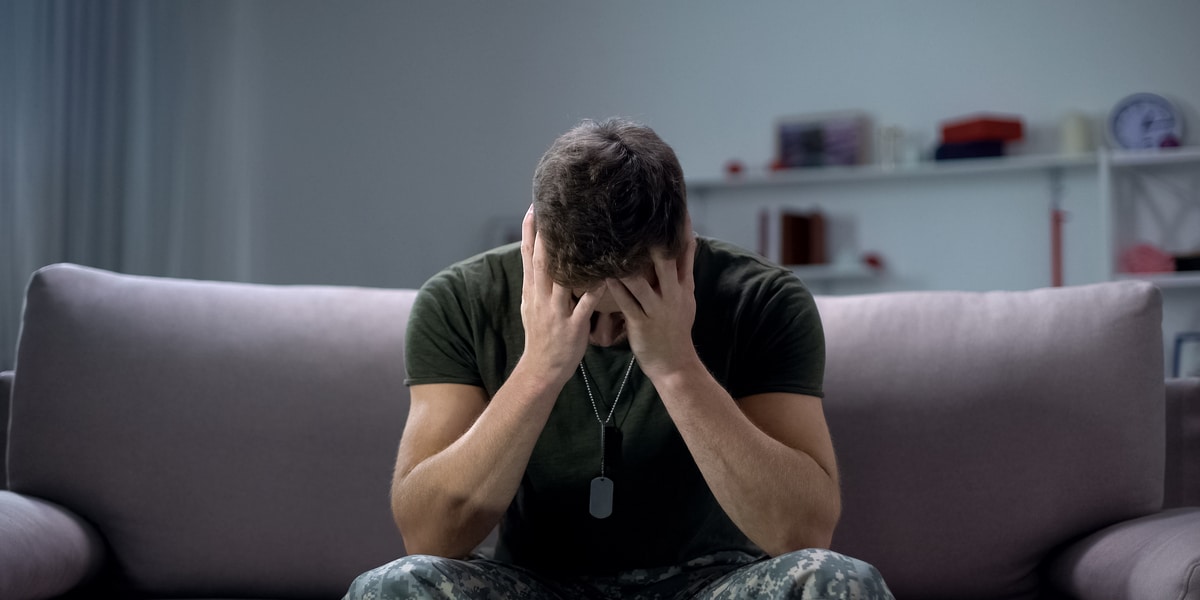 Man in military clothing seated on couch holding his head in his hands