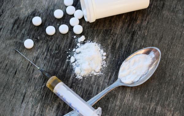White pills spilled on a table beside a pile of white powder, a teaspoon full of white powder, and a syringe full of a brown liquid