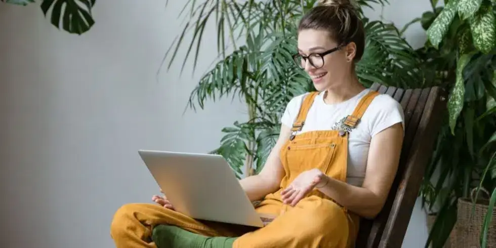 A smiling woman in overalls sitting on a chair, using a laptop