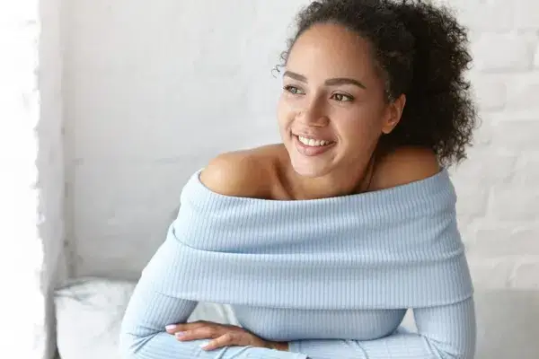 An attractive woman with curly hair sits on a bed, radiating happiness through her smile