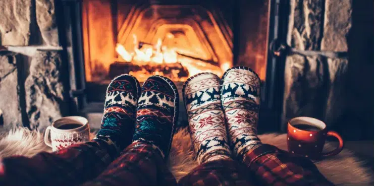Two people relax by a fireplace, wearing cozy socks and with mugs beside their feet