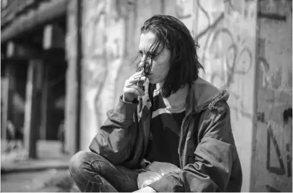 A young man smoking a cigarette in front of a graffiti covered wall, depicting an addicted person