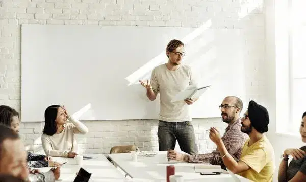 A man presenting to a group, standing behind a whiteboard, engaging in a discussion