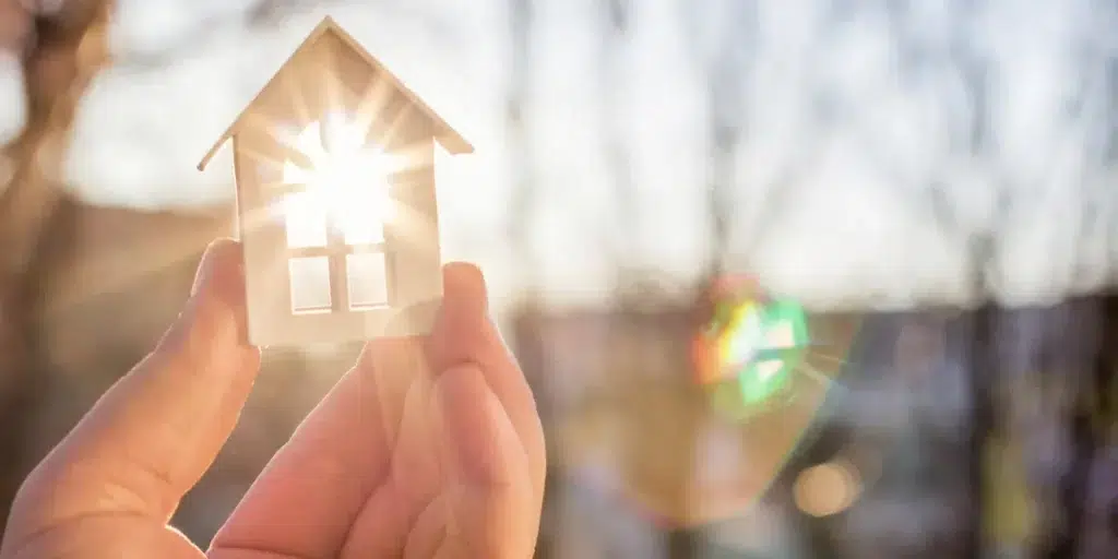 A hand holds a paper house in front of a bright sun, creating a symbolic representation of shelter and warmth