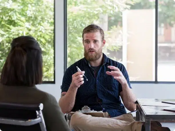 A man in a chair talking to a woman, holding a pen and gesturing